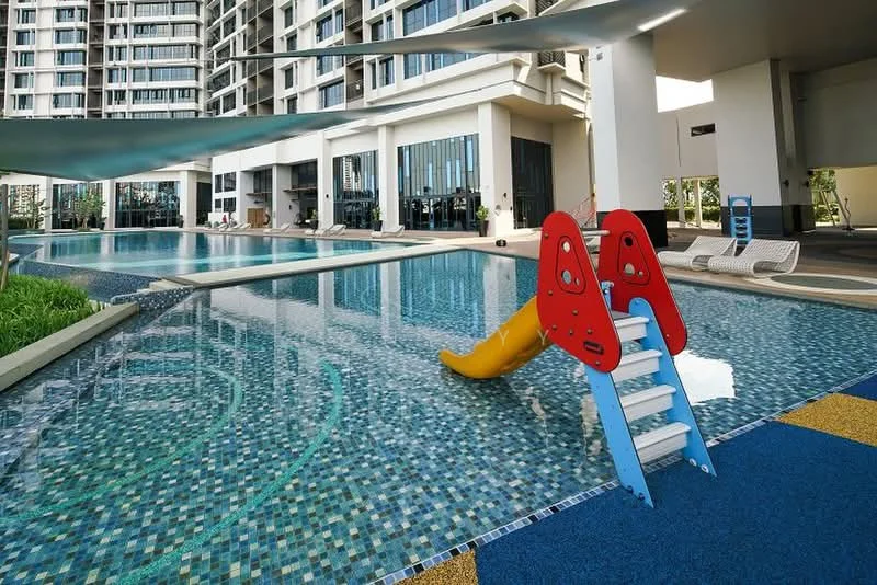 Exterior type: swimming pool deck; features: infinity pool, children's slide, pool deck with loungers, tiled pool surface, shaded seating area, landscaped greenery, blue rubber flooring, modern architecture, glass facade of building in background; style: modern.