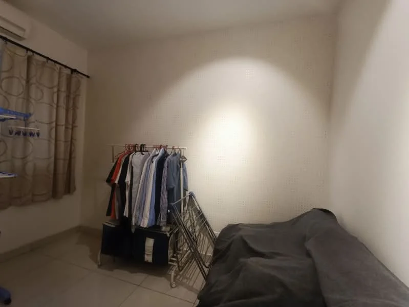 Room features: light-colored walls, tiled flooring, curtains with circular pattern, clothes rack with hanging clothes, folding drying rack, sofa covered with dark fabric, wall-mounted air conditioning unit, soft lighting from wall.