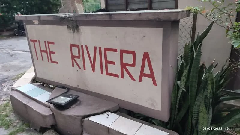 Exterior features: signage with 'THE RIVIERA' in red letters, concrete base, surrounding greenery, paved walkway, fence in background.