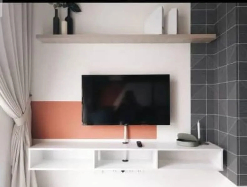 Living features: wall-mounted TV, white TV console, grey tiled accent wall, orange painted wall section, floating shelf above TV, decorative items on shelf, curtains, remote control on console; style: modern.