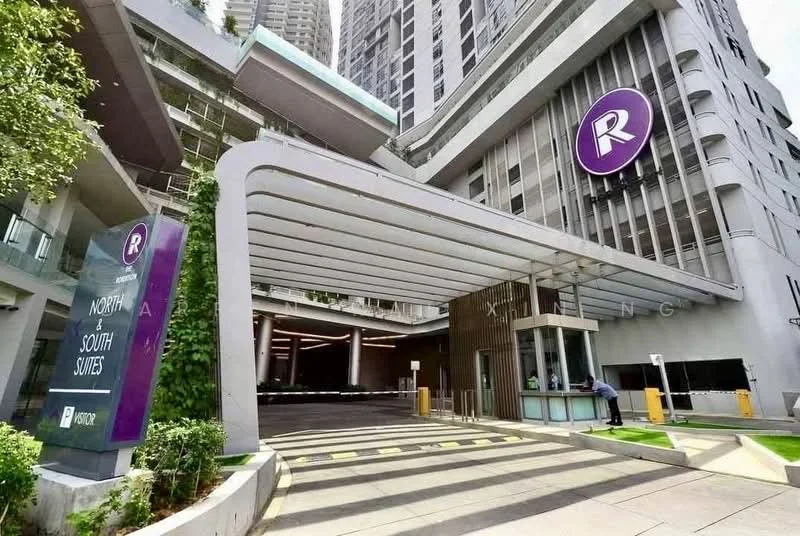 Exterior type: high-rise condominium; features: modern facade with glass and concrete, covered entrance with awning, landscaped greenery at entrance, signage with logo, security guard station, driveway with interlocking pavers, multiple storeys visible, surrounded by urban environment; style: modern.