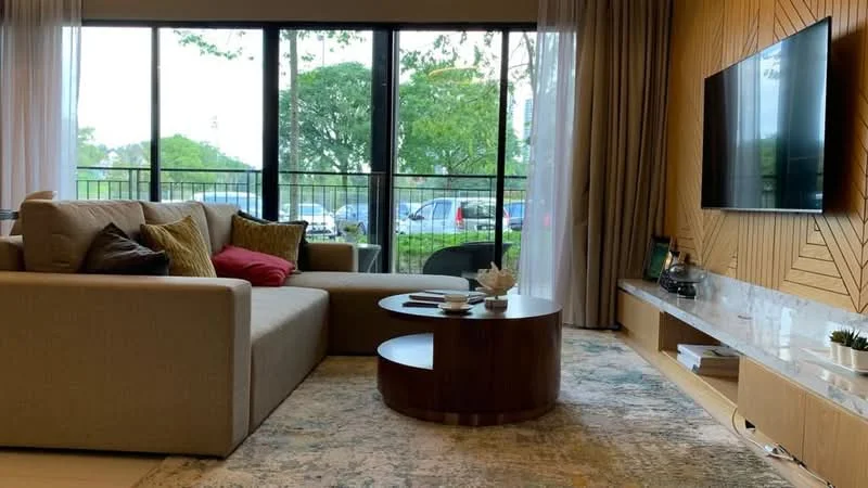 Living features: L-shape sectional sofa in beige, decorative cushions in various colors, round coffee table with wooden base, area rug with geometric pattern, wall-mounted TV, wooden TV console, large floor-to-ceiling windows, sheer curtains, wood panel accent wall, indoor plants, decorative items on console; style: modern; view: garden.