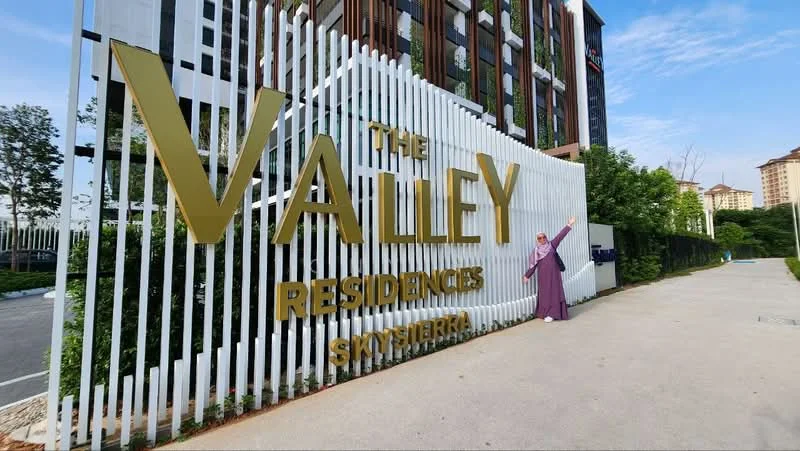 Exterior type: residential development; features: large golden signage with 'THE VALLEY RESIDENCES', white vertical slatted fencing, landscaped greenery, paved walkway, surrounding trees, modern architectural design, clear blue sky; style: modern.