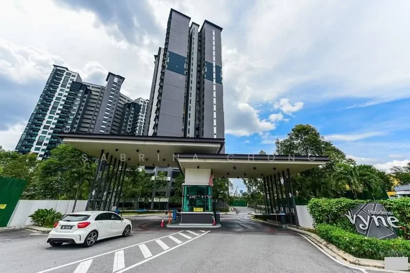 Exterior type: high-rise condominium; features: modern facade with grey and black cladding, double-volume main lobby, guard house at entrance, landscaped greenery, paved driveway, visible parking area, surrounded by mature trees, signage for 'Residences Vyne'; style: modern.