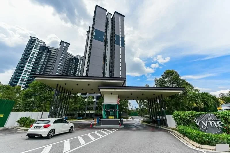 Exterior type: high-rise condominium; features: modern facade with grey cladding, multiple storeys, guard house at entrance, landscaped greenery, paved driveway, security gate, surrounded by mature trees, visible parking area; style: modern.