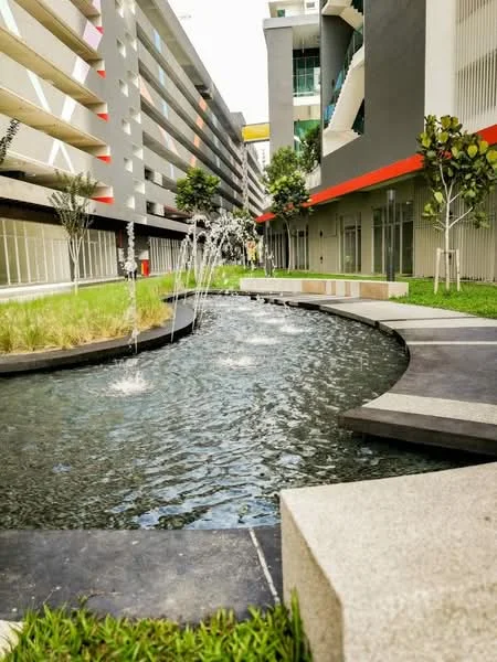 Exterior type: landscape garden; features: water feature with fountain, curved stone pathway, manicured lawn, paved seating area, surrounding greenery with trees, modern architectural backdrop, concrete planters; style: modern.