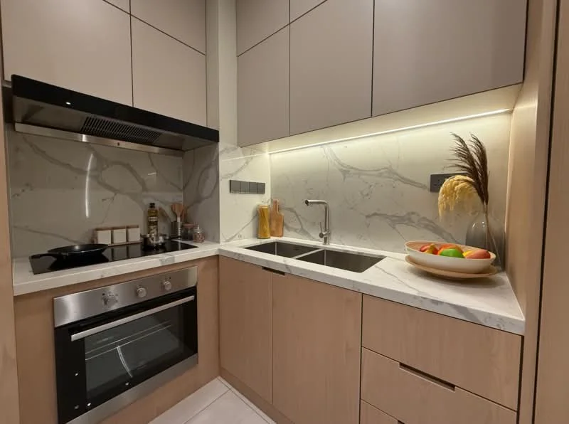 Kitchen features: light wood cabinetry, grey upper cabinets, white marble countertop, double-bowl stainless-steel sink, chrome pull-down faucet, built-in oven, 4-burner gas hob, marble backsplash, under-cabinet lighting, fruit bowl with apples and oranges, kitchen utensils on countertop; style: modern.