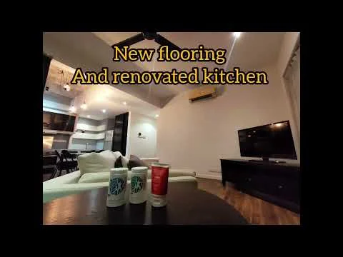 Living features: new wooden flooring, white painted walls, exposed beams on ceiling, 5-blade ceiling fan, wall-mounted TV, dark wood TV console, sofa in light color, decorative items on coffee table, air conditioning unit; style: modern. Kitchen features: renovated kitchen, open layout, kitchen island, modern cabinetry, stainless-steel appliances; style: modern.