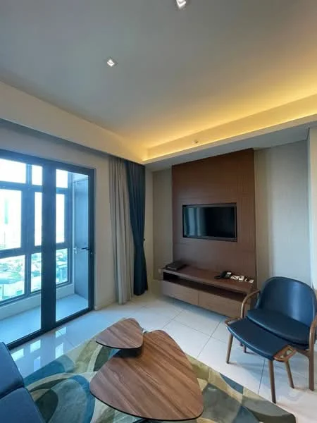 Living features: floor-to-ceiling windows, grey curtains, wooden accent wall, wall-mounted TV, light wood coffee table, blue upholstered chair, recessed downlights, white painted walls, tile flooring, sofa with patterned upholstery, decorative area rug; style: modern; view: city.