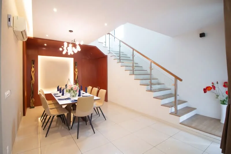 Dining features: rectangular dining table with seating for 8, upholstered dining chairs in beige, chandelier with multiple bulbs, recessed downlights, white painted walls, cream tiled flooring, wooden staircase with glass railing, decorative wall art, floor-to-ceiling windows, potted flowers in ceramic pots; style: modern.