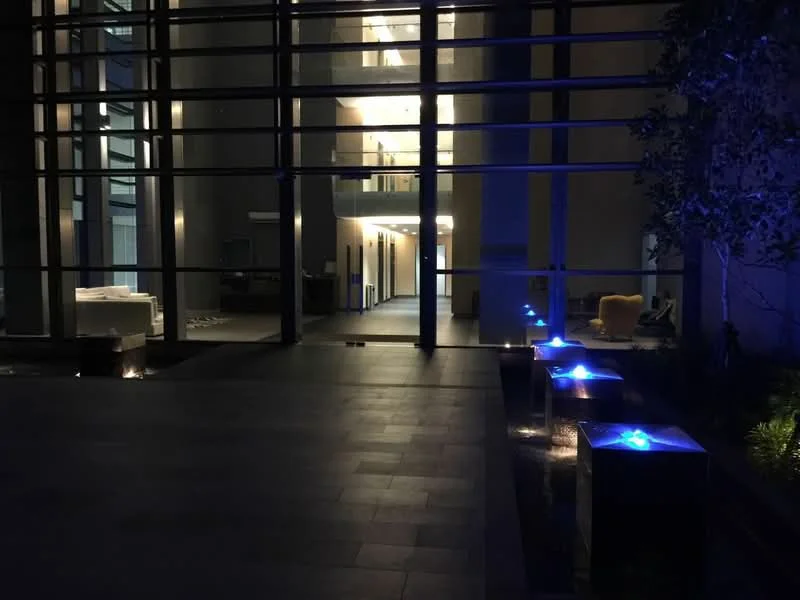 Exterior type: high-rise condominium; features: glass facade, illuminated water features, outdoor seating area, landscaped garden, stone-paved walkway, modern lighting, manicured plants; style: modern.