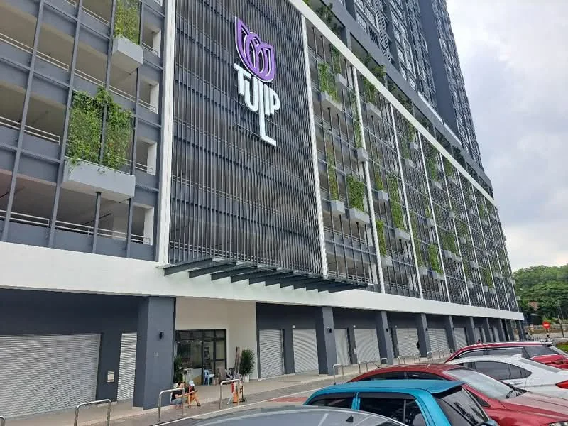 Exterior type: high-rise condominium; features: glass curtain wall, green vertical gardens, projecting balconies, signage with logo, roller shutters on ground floor, covered entrance with awning, manicured landscaping in front, open-air parking lot; style: modern.