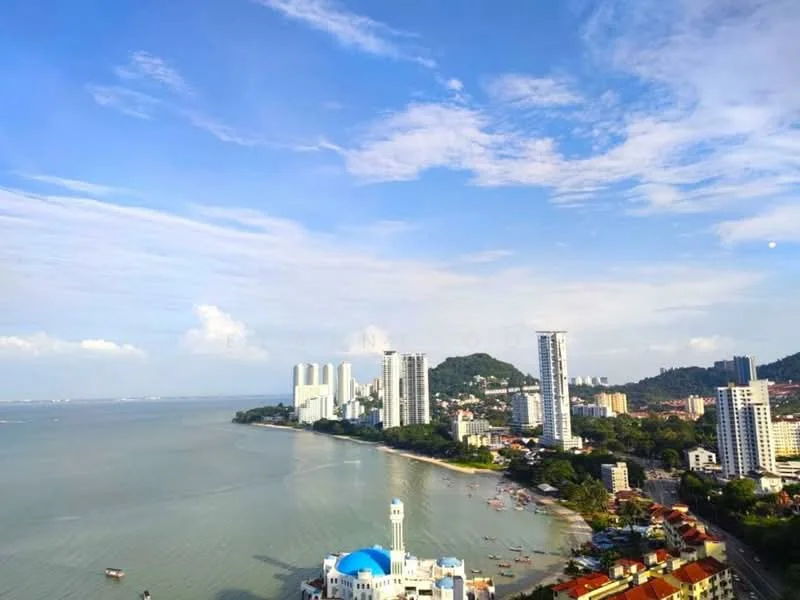 Exterior type: high-rise condominium; features: city skyline view, waterfront location, multiple high-rise buildings, coastal landscape, blue sky with clouds, beach area, boats in the water, green hills in background; style: modern.
