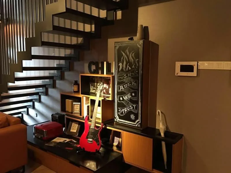 Room features: spiral staircase, wooden shelving unit, black accent wall, decorative items on shelves, red electric guitar, black refrigerator with graphic design, orange sofa, wall-mounted thermostat, floor lamp, artistic wall decor; style: modern.