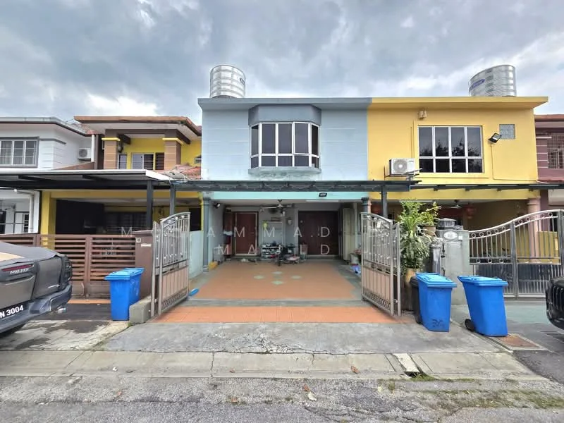 Exterior type: linked terrace house; features: two-storey building, blue and yellow painted facade, front porch with tiled flooring, two water tanks on the roof, metal gate at entrance, driveway with interlocking pavers, two blue garbage bins, windows with grilles, covered carport, surrounded by residential buildings; style: modern.