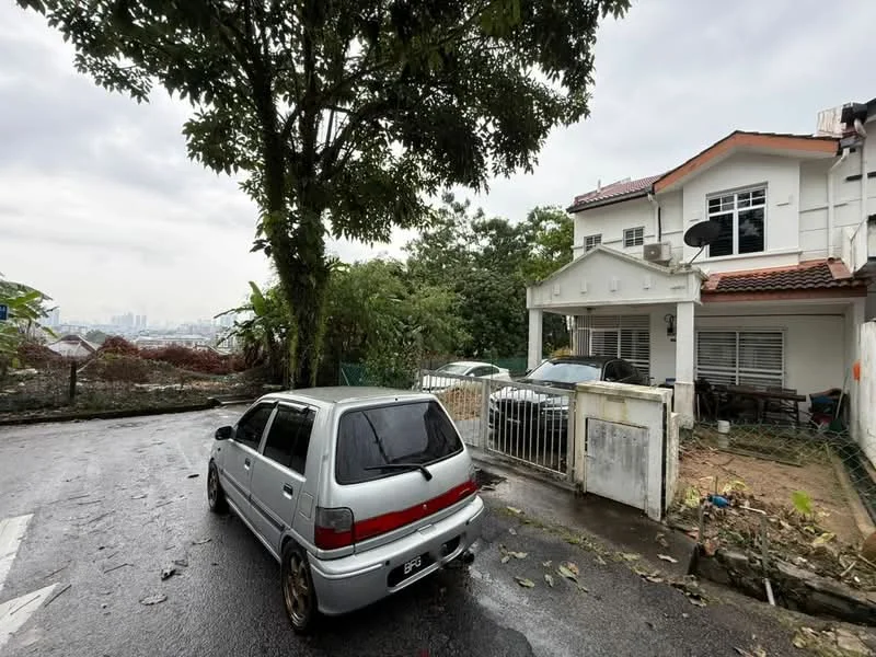 Exterior type: detached bungalow; features: white painted facade, red tiled roof, two-storey structure, front porch with railing, garden area with overgrown vegetation, driveway with parked cars, surrounded by trees, city skyline visible in background; style: modern.