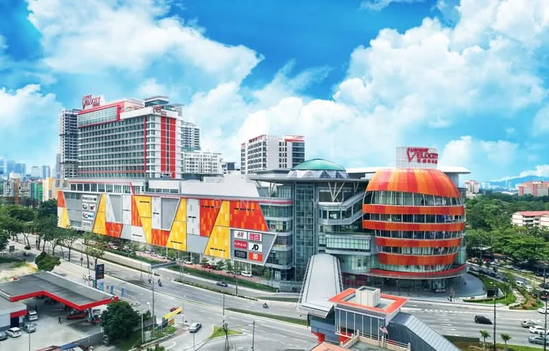 Exterior type: shopping mall; features: colorful facade with orange and yellow accents, glass dome structure, multiple levels, large signage on the building, surrounded by landscaped greenery, adjacent to a main road, parking area visible in the foreground; style: modern.