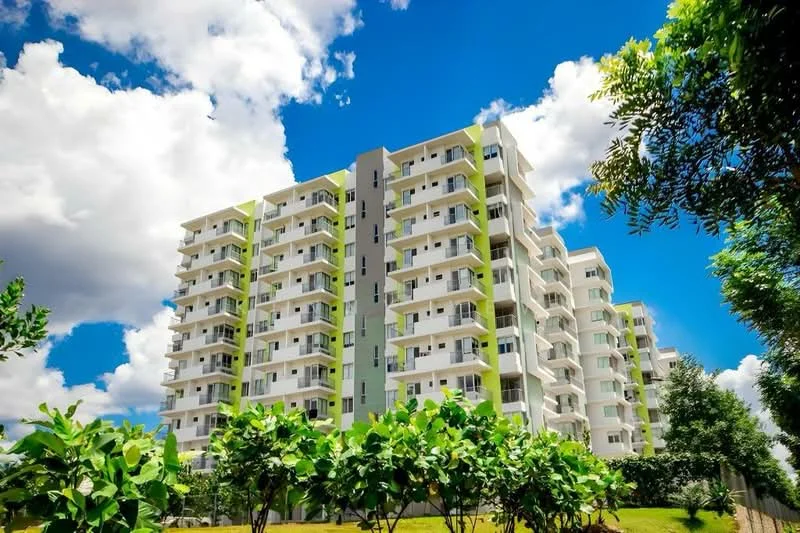 Exterior type: mid-rise condominium; features: white painted facade, green accent panels, balconies with glass railings, multiple storeys, landscaped garden area, mature trees, manicured lawn, clear blue sky, clouds, surrounded by greenery; style: modern.