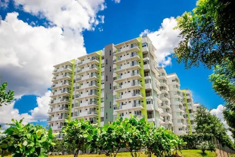 Exterior type: mid-rise condominium; features: green accent panels on facade, balconies on each floor, white painted facade, landscaped garden in foreground, mature trees surrounding the building, clear blue sky with clouds, multiple storeys, modern architectural design; style: modern.