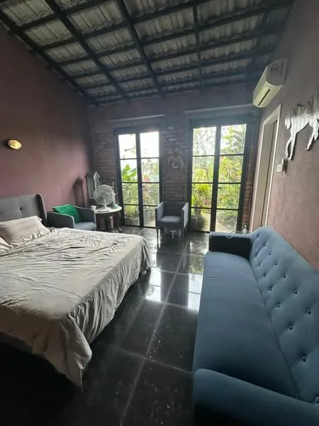 Bedroom features: king-size bed with beige bedding, grey upholstered headboard, black marble flooring, exposed brick wall, dark purple painted walls, two large windows with black frames, green throw blanket on bed, two armchairs in grey fabric, small wooden side table, air conditioning unit on wall, decorative wall art; style: modern; view: garden.