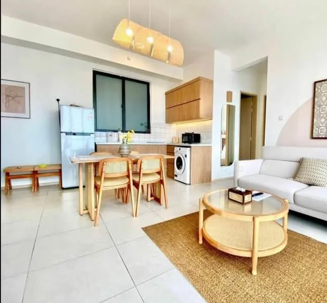 Living features: white walls, tile flooring, sofa in white fabric, coffee table with rattan base, area rug with natural fibers, pendant light fixture, dining table with wooden legs, 4 dining chairs, refrigerator in stainless steel, microwave, washing machine, window with black frame, built-in cabinetry, decorative artwork on wall; style: modern.