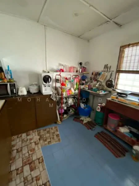 Kitchen features: white painted walls, vinyl tile flooring in blue, wood cabinetry, countertop with clutter, microwave, kettle, dish rack, sink, various kitchen utensils, window with blinds, plastic storage containers, area rug, broom and dustpan.