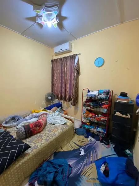 Bedroom features: yellow painted walls, wall-mounted split AC, ceiling fan with light, double bed with patterned bedding, bedside pillows, window with curtain, clothes rack, black dresser, carpet with abstract pattern, scattered clothes on the floor.