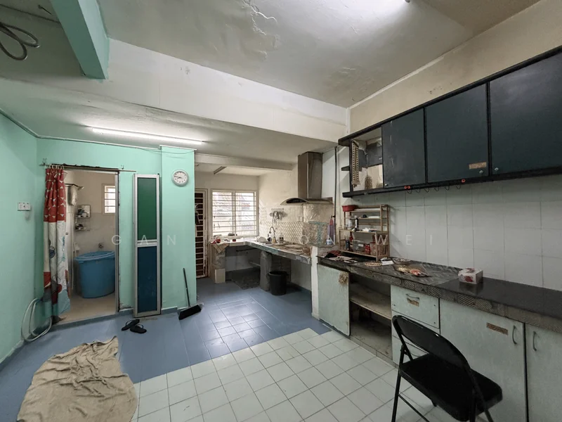 Kitchen features: green painted walls, white tiled walls, blue vinyl tile flooring, stainless-steel countertop, built-in cabinets with blue doors, single-bowl stainless-steel sink, chrome pull-down faucet, freestanding stove, exhaust hood, wall-mounted clock, window with metal grilles, open shelving, plastic chair, cleaning cloth on floor.
