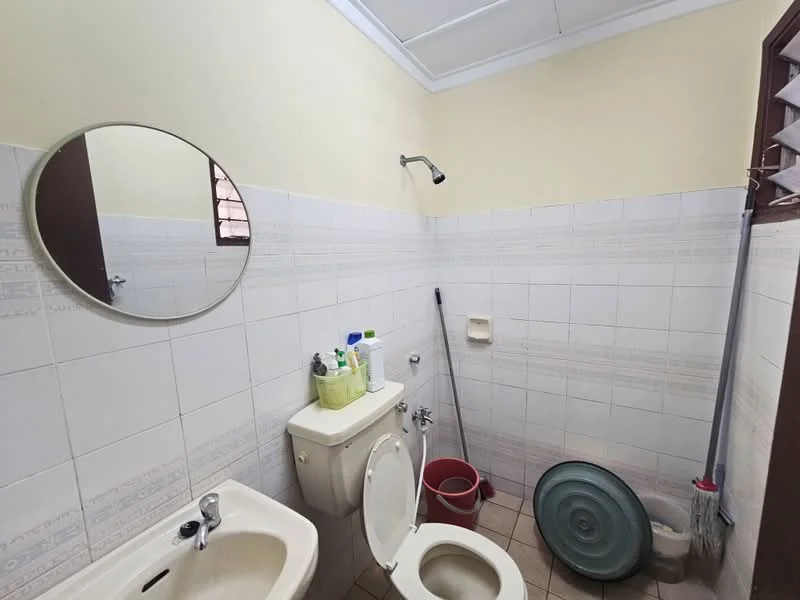 Bathroom features: white tiled walls, light beige tiled flooring, round mirror, single-sink vanity, one-piece toilet, shower head with wall mount, soap dispenser, toilet brush, bucket, broom, cleaning supplies.