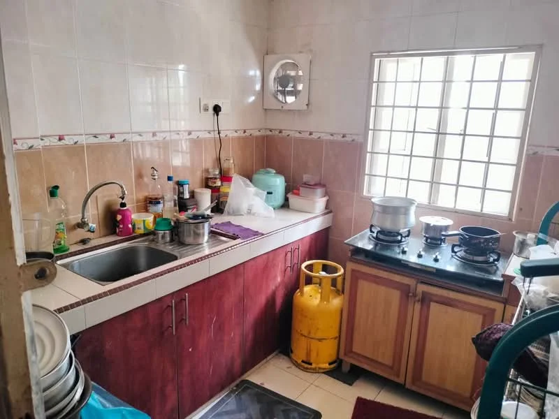 Kitchen features: single-bowl stainless-steel sink, chrome faucet, wood cabinetry in dark brown, white tiled walls, cream tiled backsplash, gas stove with two burners, metal cooking pots, plastic storage containers, green gas cylinder, window with grid pattern, floor mat in dark color, countertop with cluttered items.