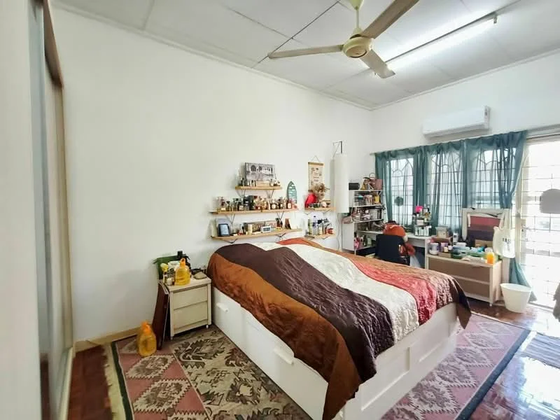 Bedroom features: queen-size bed with patterned bedding, wooden flooring, white painted walls, ceiling fan, air conditioning unit, green curtains, shelves with decorative items, nightstand with lamp, dressing table, various personal items on surfaces, area rug with geometric pattern, large windows with grid design.