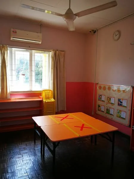Room features: pink painted walls, orange accent wall, window with sheer curtains, 1 wall-mounted split AC, ceiling fan, wooden shelving unit, orange table with markings, yellow plastic chair, clock on wall, vinyl flooring.