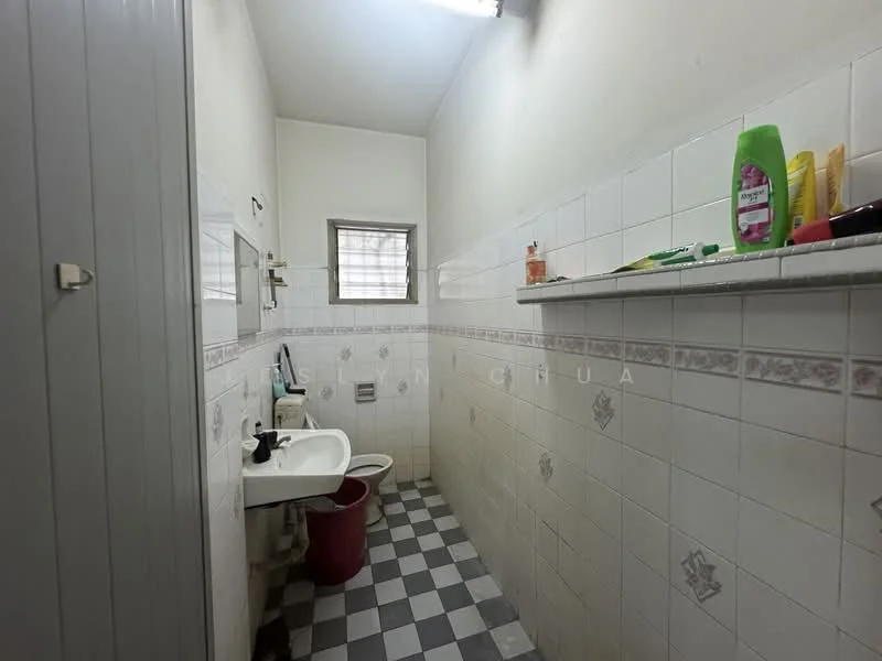 Bathroom features: white tiled walls, grey and white checkered floor tiles, single-sink vanity with mirror, toilet, window with sheer curtain, shelf with toiletries, overhead fluorescent lighting, red waste bin.