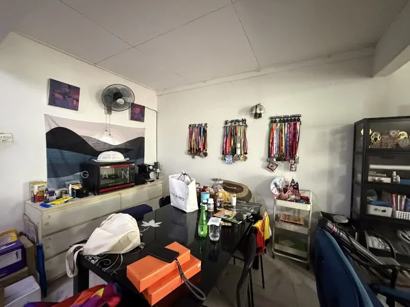 Room features: white painted walls, ceiling fan, dining table, black chairs, glass display cabinet, wall-mounted medals, decorative wall art, various personal items on table, plastic storage boxes, bottled drinks, shopping bags.