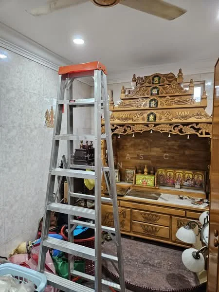 Room features: ladder, wooden altar with decorative carvings, multiple framed religious images, wall-mounted lighting fixtures, white tiled walls, light-colored ceiling with recessed lighting, storage cabinet, various items on the floor including baskets and cloth; style: traditional.