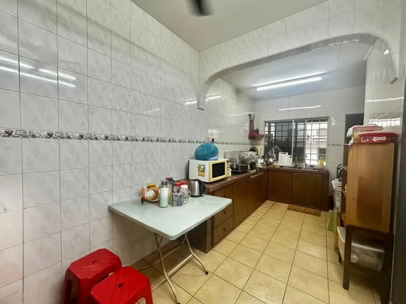 Kitchen features: white tiled walls, cream tiled backsplash, brown wooden cabinetry, white countertop, microwave, stainless-steel sink, kitchen appliances on countertop, table with white surface, two red plastic stools, tile flooring, window with black grille.