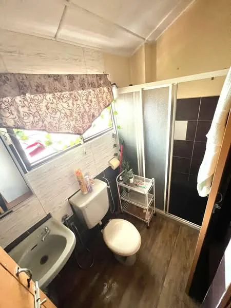 Bathroom features: white painted walls, dark tile flooring, shower enclosure with glass door, toilet, sink with chrome faucet, window with sheer curtain, small storage cart, plant decoration.