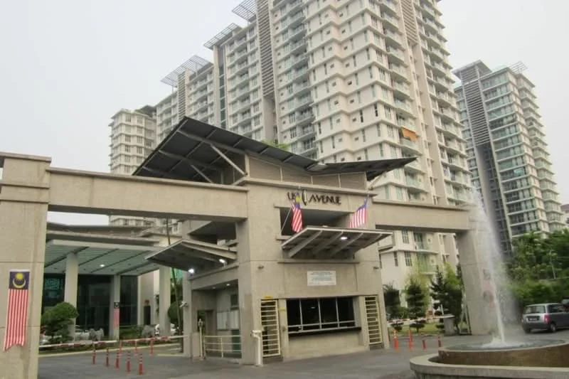 Exterior type: high-rise condominium; features: modern facade with glass and concrete, double-volume main lobby entrance, feature water wall at entrance, landscaped garden with greenery, security guard house at entrance, flag poles with Malaysian flags, visible parking area, surrounded by mature trees; style: modern.