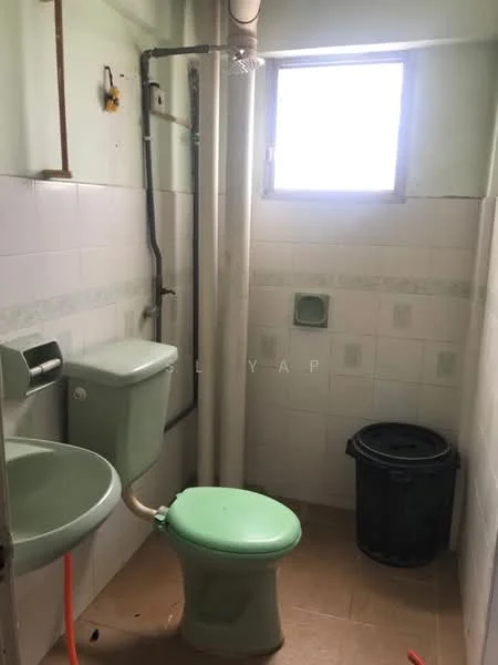 Bathroom features: green toilet, green basin, white tiled walls, light green accents, window for natural light, floor tile flooring, plumbing pipes visible, waste bin.