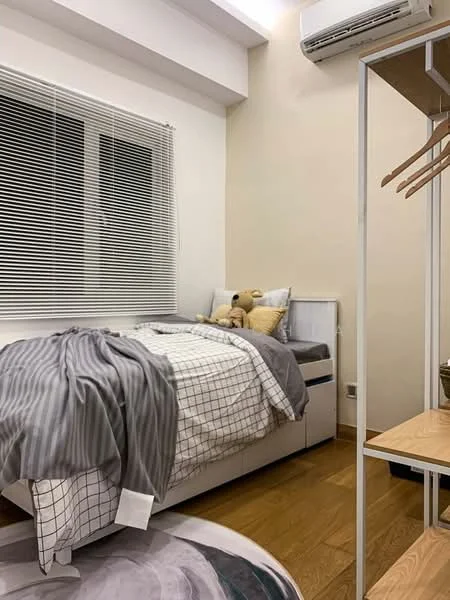 Bedroom features: single bed with white and grey bedding, plaid patterned throw blanket, stuffed teddy bear on bed, white walls, wooden flooring, floor-to-ceiling window with white blinds, air conditioning unit on wall, open wardrobe with hanging rods, wooden shelving unit, decorative area rug; style: modern.