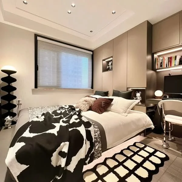 Bedroom features: queen-size bed with white bedding, black and white patterned throw blanket, black and white area rug, built-in wardrobe with sliding doors, desk with chair, wall-mounted shelving with books, floor lamp with unique design, window with blinds, recessed ceiling lights, decorative pillows, wall art, wooden flooring; style: modern.