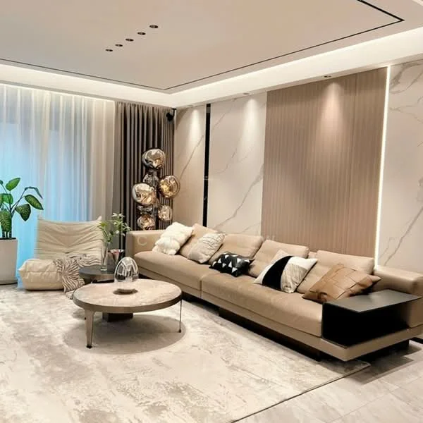 Living features: L-shape sectional sofa in beige, decorative cushions in various colors, round coffee table with marble top, area rug with neutral tones, floor-to-ceiling windows with sheer curtains, wall-mounted lighting fixtures, accent wall with vertical stripes, potted indoor plant, decorative floor lamp, recessed ceiling lights; style: modern.