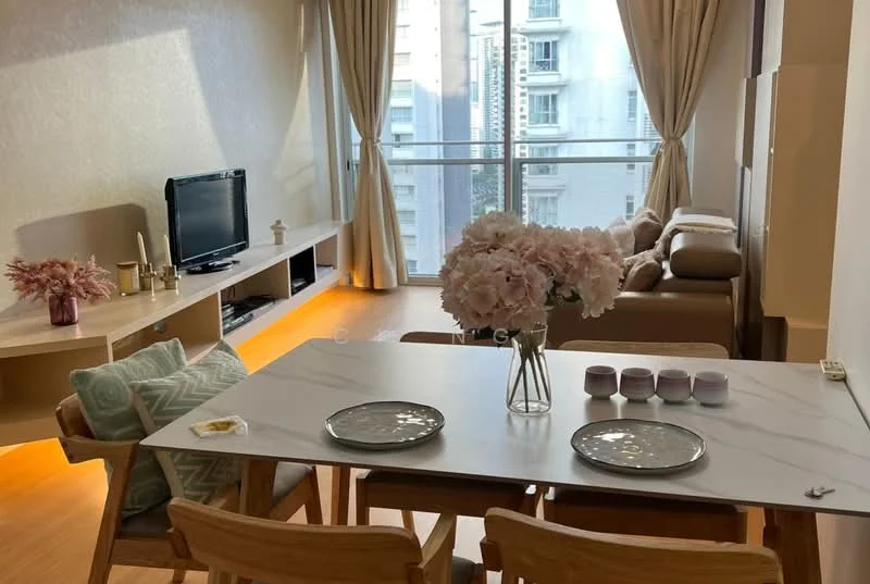 Dining features: marble dining table, 4 wooden dining chairs, decorative vase with flowers, 2 plates on the table, 3 small cups on the table, wall-mounted TV, light wood flooring, floor-to-ceiling windows with sheer curtains, sofa in the background, built-in shelving unit; style: modern; view: city.