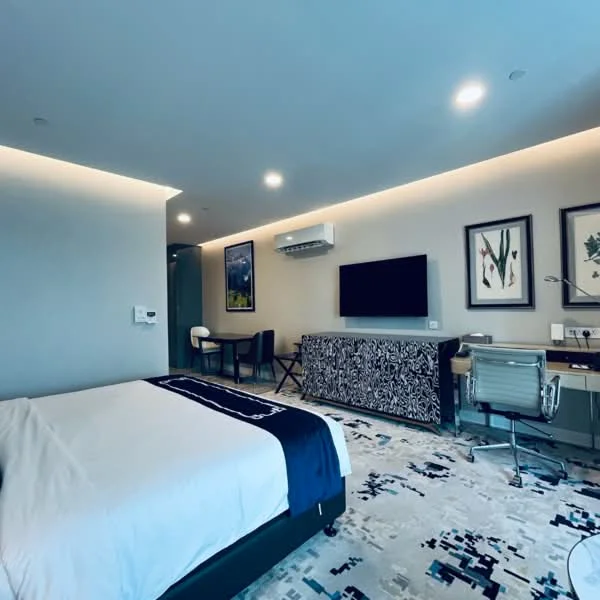 Bedroom features: king-size bed with white bedding, upholstered headboard, wall-mounted TV, air conditioning unit, desk with chair, decorative artwork on walls, carpet flooring with abstract pattern, recessed lighting, nightstand with lamp, built-in wardrobe; style: modern.
