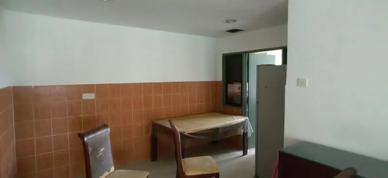 Room features: white painted walls, brown tiled wall, plain ceiling, flooring, wooden dining table, two wooden chairs, grey cabinet, window with green frame, plastic covering on table.