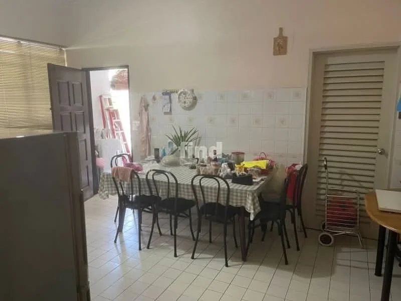 Kitchen features: checkered tablecloth on dining table, black dining chairs, white tiled walls, white tiled flooring, stainless-steel fridge, wall-mounted clock, kitchen cabinets with louvered doors, various kitchen utensils on the table, potted plant on the table, open doorway leading to another room, natural light from window, shelves with items visible.
