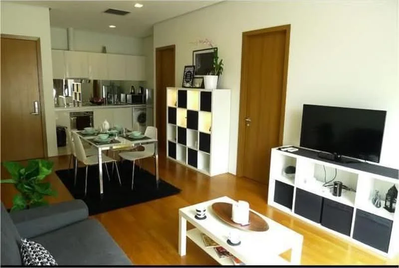 Living features: grey fabric sofa, black area rug, white coffee table, TV console with black and white storage, wall-mounted TV, decorative items on shelves, potted plant, wooden flooring, recessed lighting, open shelving unit, dining table with four chairs, kitchen area visible in background; style: modern.