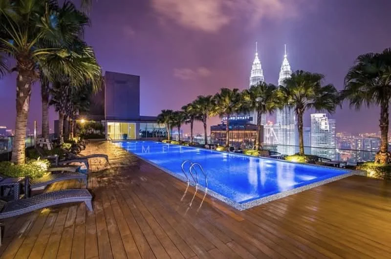 Exterior type: high-rise condominium; features: infinity pool, pool deck with loungers, tropical landscaping with palm trees, wooden decking, city skyline view, night lighting, surrounded by greenery; style: modern.