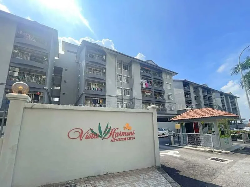 Exterior type: low-rise apartment block; features: grey painted facade, balconies with railings, visible laundry on balconies, paved driveway, landscaped entrance with signage, gated entry with guardhouse, parking area, surrounded by palm trees; style: modern.
