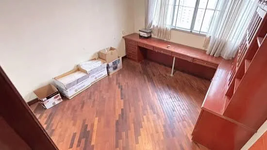 Study features: wood flooring, large window with sheer curtains, built-in wooden desk, wooden cabinetry, boxes stacked against the wall, neutral wall color, natural light from window, open layout; style: modern; view: city.